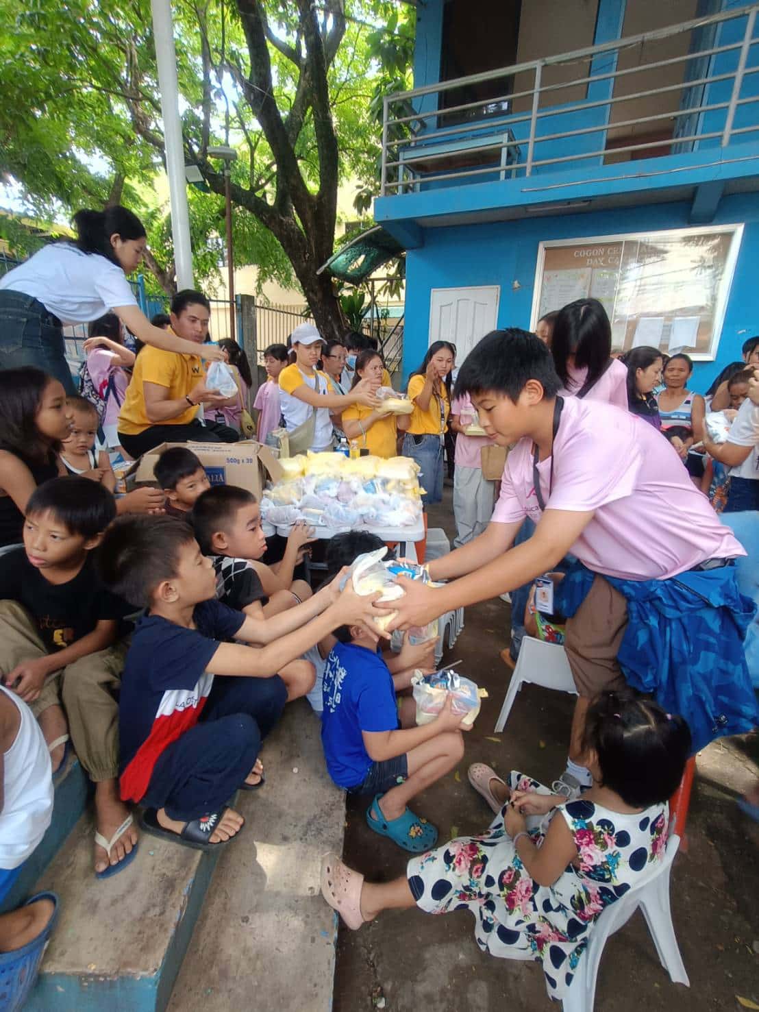 學生站在椅子上將食物遞給坐在地上的當地孩子
